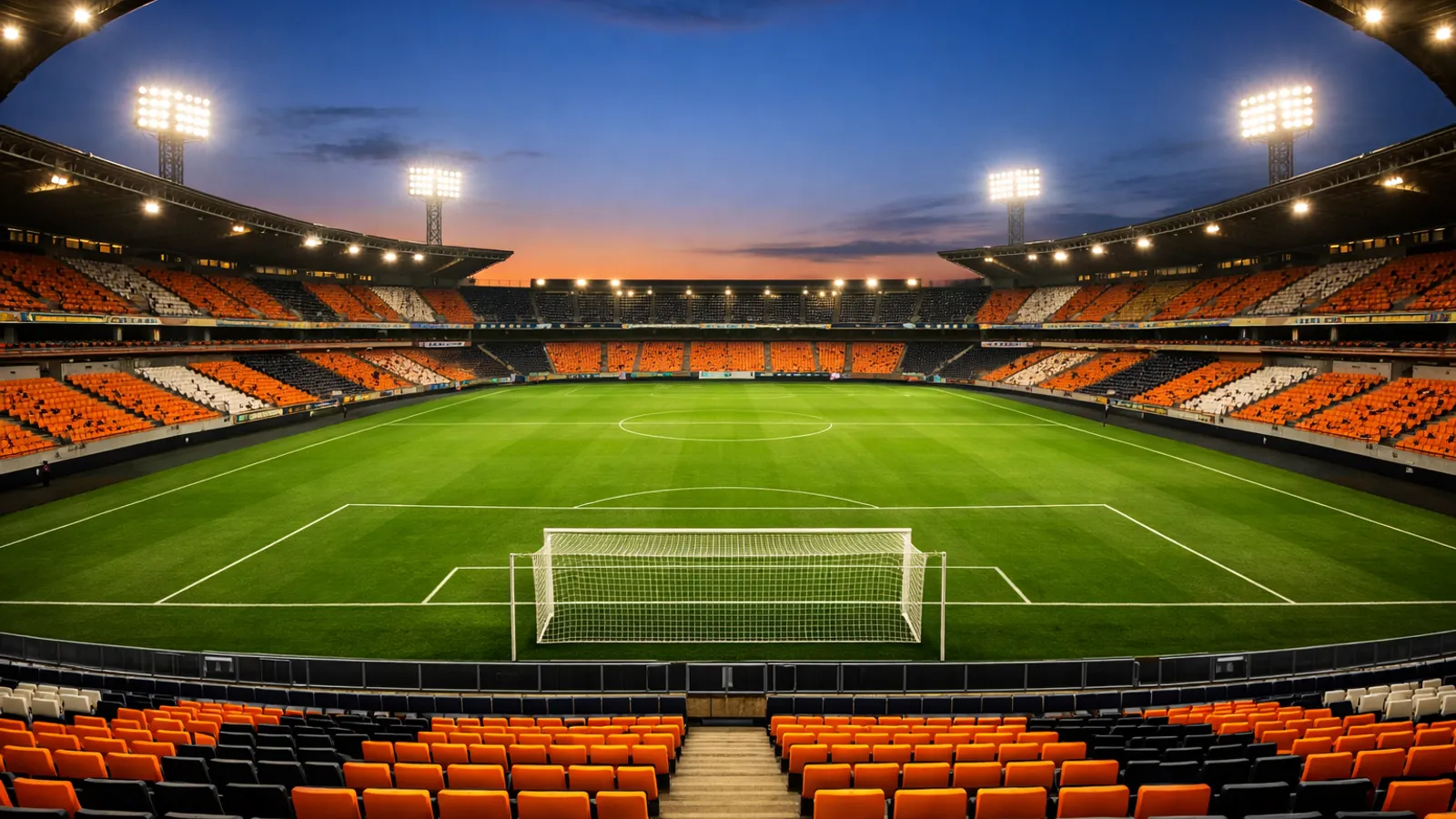 Fotografia realista ampla de estádio de futebol vazio visto da arquibancada, gramado verde e refletores acesos no céu azul do entardecer.